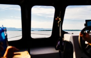 L’image montre une vue pittoresque depuis l’intérieur d’un véhicule, probablement un bateau, avec une fenêtre encadrant un magnifique paysage d’eau et de montagnes au loin. L’intérieur est légèrement flou, mais on peut voir un volant à droite et des mains tenant une carte à gauche. La lumière vive suggère qu’il fait jour, donnant une impression de voyage ou d’exploration.